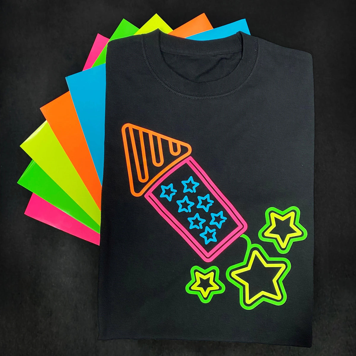 Siser Easy Glow HTV in five neon colors, fanned behind a black tee with a heat pressed neon firework and stars design